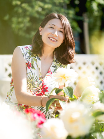 Wide smiling woman is admiring of blooming roses in public park. summer vibes. Tropical plants and flowers in bloom in garden.の写真素材