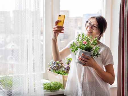 Woman is shooting selfie with blooming carnation flower. Houseplants and microgreens on windowsill. Growing edible organic basil, arugula for healthy nutrition. gardening at home.の写真素材
