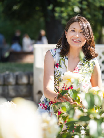 Caucasian woman is admiring of blooming roses in public park. summer vibes. Tropical plants and flowers in bloom in garden.の写真素材