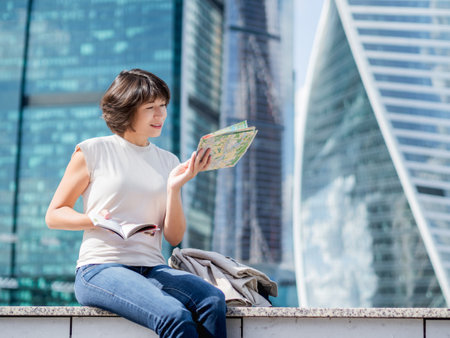 Smiling woman is reading paper map sitting on background of buildings with glass walls. Travel around city. urban tourism. Modern architectural landmarks.の写真素材