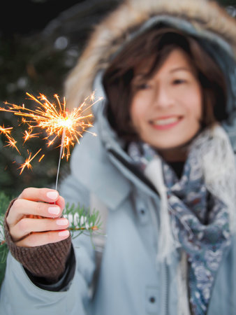 Smiling woman celebrates Christmas or New Year with bright sparkler. Bengal fire, traditional firework for winter holiday celebration outdoors.の写真素材