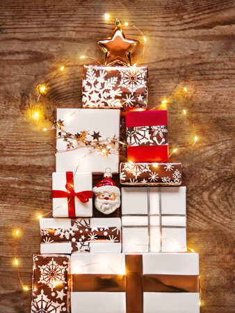 New Year presents in shape of Christmas tree on shabby wooden background. Gifts in white and golden paper with bright ribbons and light bulbs.の写真素材