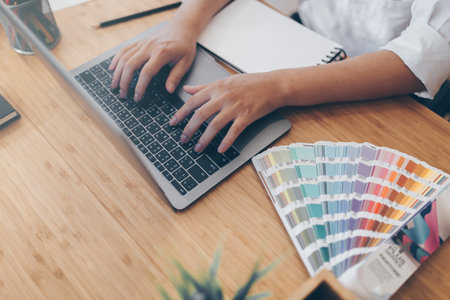 A man hands is working. One hand is taking on laptop and keep a pen writing on notebook. He working on wood table cream colour. He is designer and he wearing white shirts.の写真素材