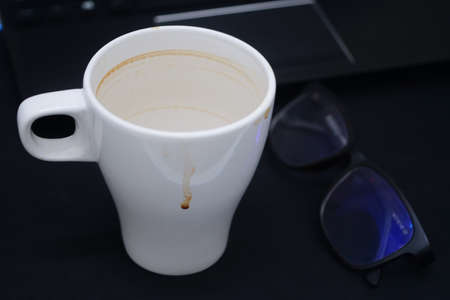 Empty white cup of coffee with coffee stain near a laptop and glasses. Dark backgroundの写真素材