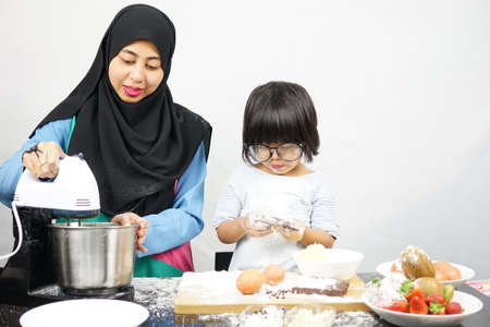 Asian Muslim mother and cute daughter preparing the dough to make a cake in kitchen room at home. Messy kitchen. Motherhood concept. Happy Familyの写真素材