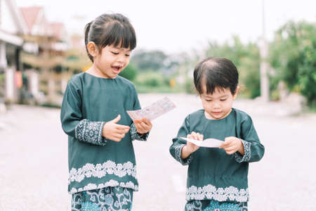Cute Malay girls siblings sisters in Malay traditional cloth showing his happy reaction after received money pocket during Eid Fitri or Hari Raya celebrationの写真素材