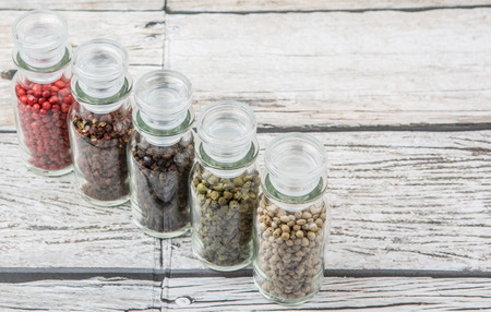 Szechuan peppercorns, green peppercorns, white peppercorns, black peppercorns and pink peppercorns in glass vial over wooden backgroundの写真素材