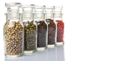 Szechuan peppercorns, green peppercorns, white peppercorns, black peppercorns and pink peppercorns in glass vial over white backgroundの写真素材