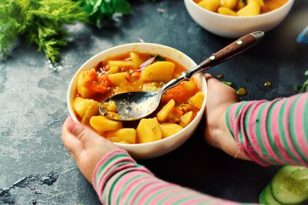 Photo of food on a dark background. Childrens hands and food. The child holds hands a bowl with vegetable stew. Baby is having lunch. Food on a concrete background.の写真素材