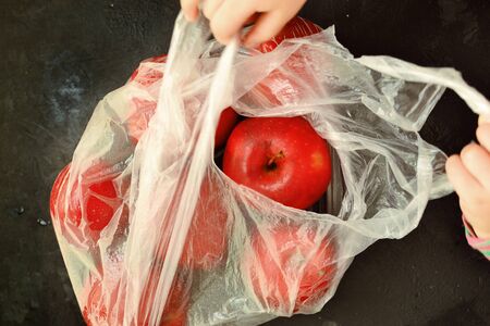 Red ripe apples in a plastic bag. Dark background, top view. Buying fruit. Hands of a child in the...
