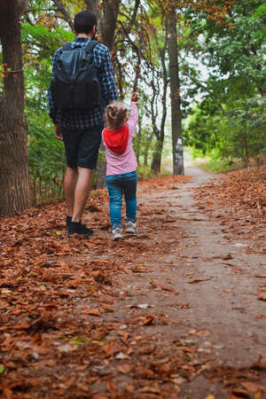 The baby goes by the hand with dad outdoors on an autumn day. View from the back. Happy family and childhood moments. Dad and child are walking in the autumn park.の写真素材