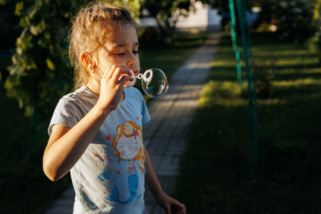 Girl 5-6 years with dark hair blowing bubbles outdoors at sunset. High quality photo