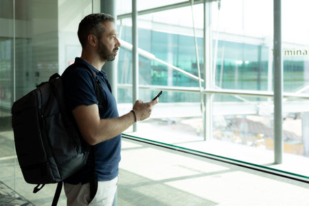 Man with bread with backpack using Phone while standing against window in airport terminal. High quality photoの写真素材