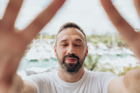 Smiling man in a white T-shirt with beard and closed eyes takes selfie on a yachts view. High quality photoの写真素材