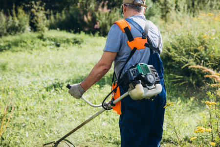 Behind the house, a man mows the grass with a trimmer on a hot, clear day. A man mows tall grass with a trimmer that has overgrown the lawn behind the house. Grass mowing concept. High quality photoの写真素材