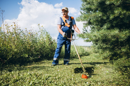 Senior man mowing grass with a hand-held trimmer in a field through the tree. High quality photoの写真素材
