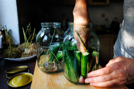 Cucumbers are being carefully packed into glass jars along with herbs in a cozy kitchen. Natural light filters in, highlighting the vibrant green produce and wooden surface.の写真素材