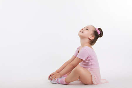 Little happy girl gymnast sitting on the floor holding his leg. The girl looks up. Light background and empty space for textの写真素材