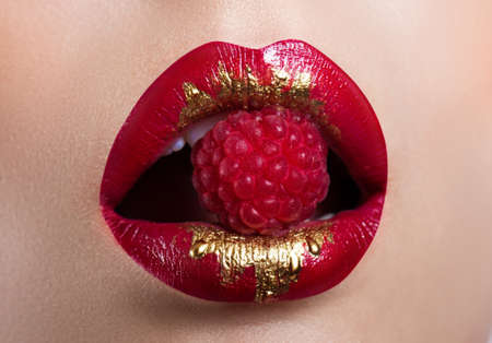Beautiful female lips closeup. Red lipstick with gold. Berry raspberry in mouthの写真素材