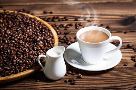 Coffee beans on a large plate and a cup of fragrant hot coffee with milk on a wooden table.の写真素材