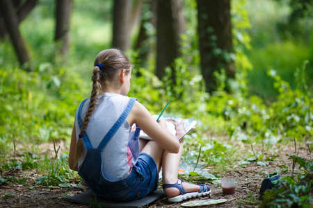 Girl painter paints trees in the park. Girl in denim overalls sitting with her backの写真素材