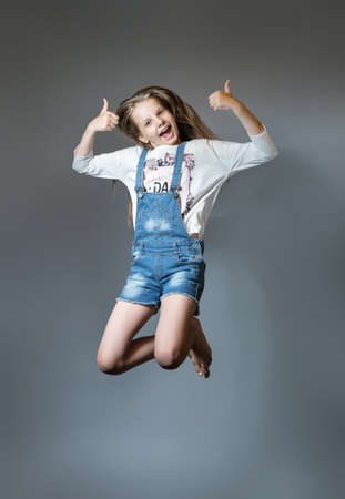 Lovely girl joyfully jumping. Cheerful teen girl in denim overalls on gray backgroundの写真素材