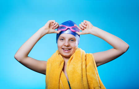 Girl swimmer in a bathing cap and glasses. The girl holds glasses for diving. She laughs. The concept of success. Yellow towel on his shoulders. Bright blue background. Horizontal photoの写真素材