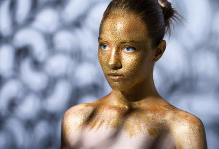 portrait of a beautiful young girl. Face painted with gold paint with glitter. abstract shadows on the background and on the model.の写真素材