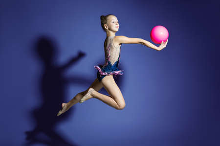 girl gymnast performs a jump with the ball. Frozen motion. Violet background. A child in a bathing suit for rhythmic gymnastics.の写真素材