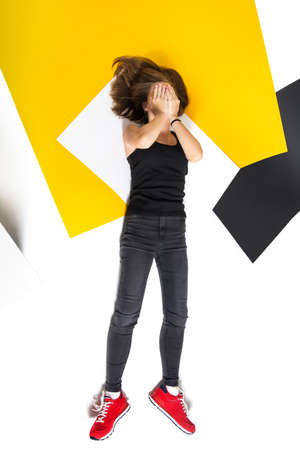 Teen girl lies covering her face with her hands. View from above. Dark clothes, red shoes. white,...