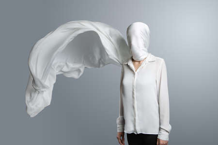 Anonymous model, covered with a white cloth. Woman in white shirt with head wrapped in waving white veil on light grey backgroundの写真素材