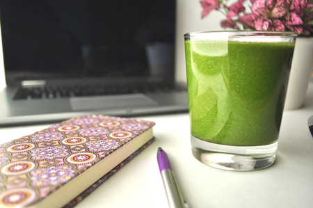 A glass of fresh green smoothie on a table with laptop computer, pink notebook, pen and a pink potted plant - a work from home workstationの写真素材