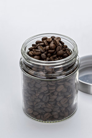 A glass jar filled with aromatic coffee beans, with metal lid placed beside it on white background. image captures rich texture and dark color of beansの写真素材
