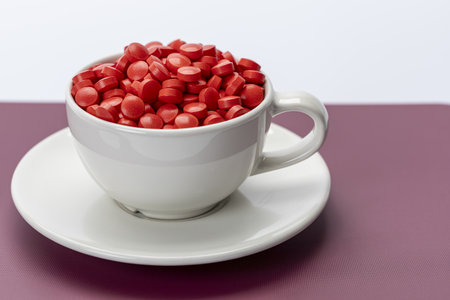 White ceramic cup filled with many red pills on saucer, concept of daily medicine, pharmaceutical healthcare, symbolic lifestyle image.の写真素材