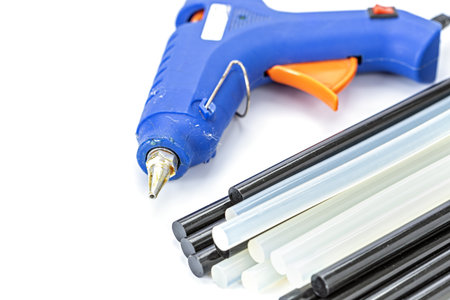 A blue hot glue gun placed beside black and white glue sticks on a white background, showing essential crafting and DIY repair tools used for bonding materials together.の写真素材