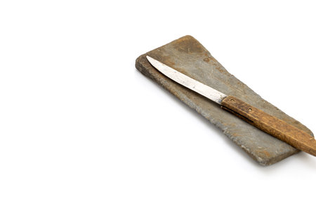 A knife with a wooden handle lays atop a gray sharpening stone, angled in the top right corner of the frame. The objects are isolated against a stark white background. Rustic, simple.の写真素材
