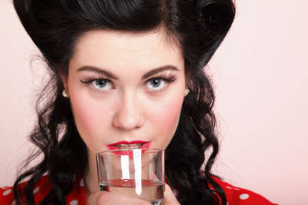 Beautiful young woman with pinup makeup and hairstyle drinking water pink backgroundの写真素材