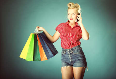 Retro style. Young woman in red dotted shirt and shorts with colourful shopping bags talking on cell...