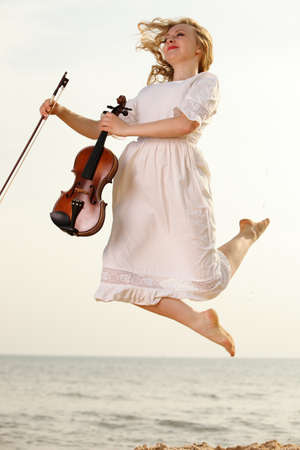Happy crazy blonde girl music lover on beach jumping with a violin. Love of music concept.の写真素材