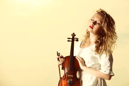 The blonde girl music lover on pier with a violin at sunset or sunrise.  Love of music concept.