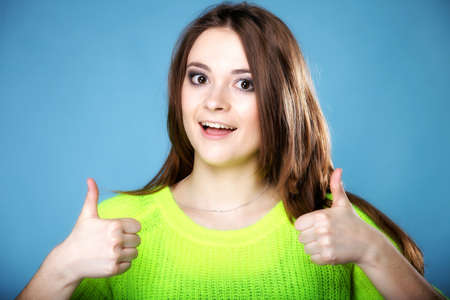 Happy smiling woman in bright vivid colour sweater with thumbs up gesture, on blue backgroundの写真素材