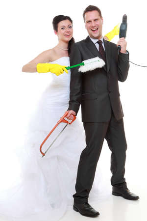 Humorous funny young couple bride groom in domestic role, sharing household chores. Woman with gloves holding duster man with drill isolated on white backgroundの写真素材