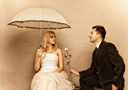 Wedding day. Portrait of romantic married couple blonde bride with umbrella and enamored groom giving a rose to girl. Studio shot sepia color, vintage photoの写真素材