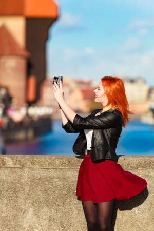 Female photographer taking pictures. Traveler woman red haired fashion girl with camera outdoors in european city, old town Gdansk river Motlawa in the background, Poland Europeの写真素材