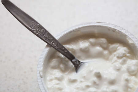 Close-up of white cottage cheese in plastic container and teaspoon on kitchen table background....