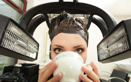 Young woman drinking coffee tea in hairdressing beauty salon. Girl dying colouring hair by hairdresser hairstylist. Modern equipment.の写真素材