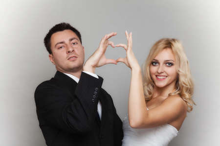 Wedding day. Happy blonde bride and groom showing making heart shape sign with hands gray backgroundの写真素材