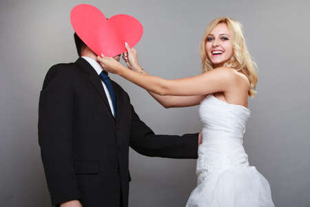 Wedding day. Happy funny blonde bride covered groom face with red heart gray backgroundの写真素材