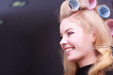 Beautiful young woman in beauty salon. Happy blond girl with hair curlers rollers by hairdresser. Hairstyle.の写真素材
