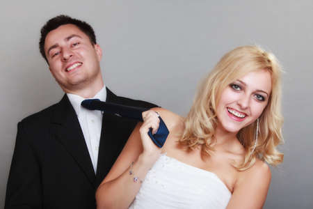 Wedding day. Portrait of happy married couple blonde bride and groom. Woman pulling on mans tie, studio shot on gray backgroundの写真素材